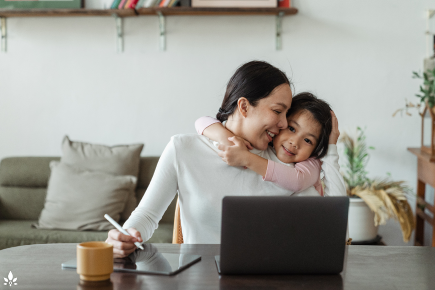 A woman hugs her daughter while working on her laptop. Attempt at work-life balance.